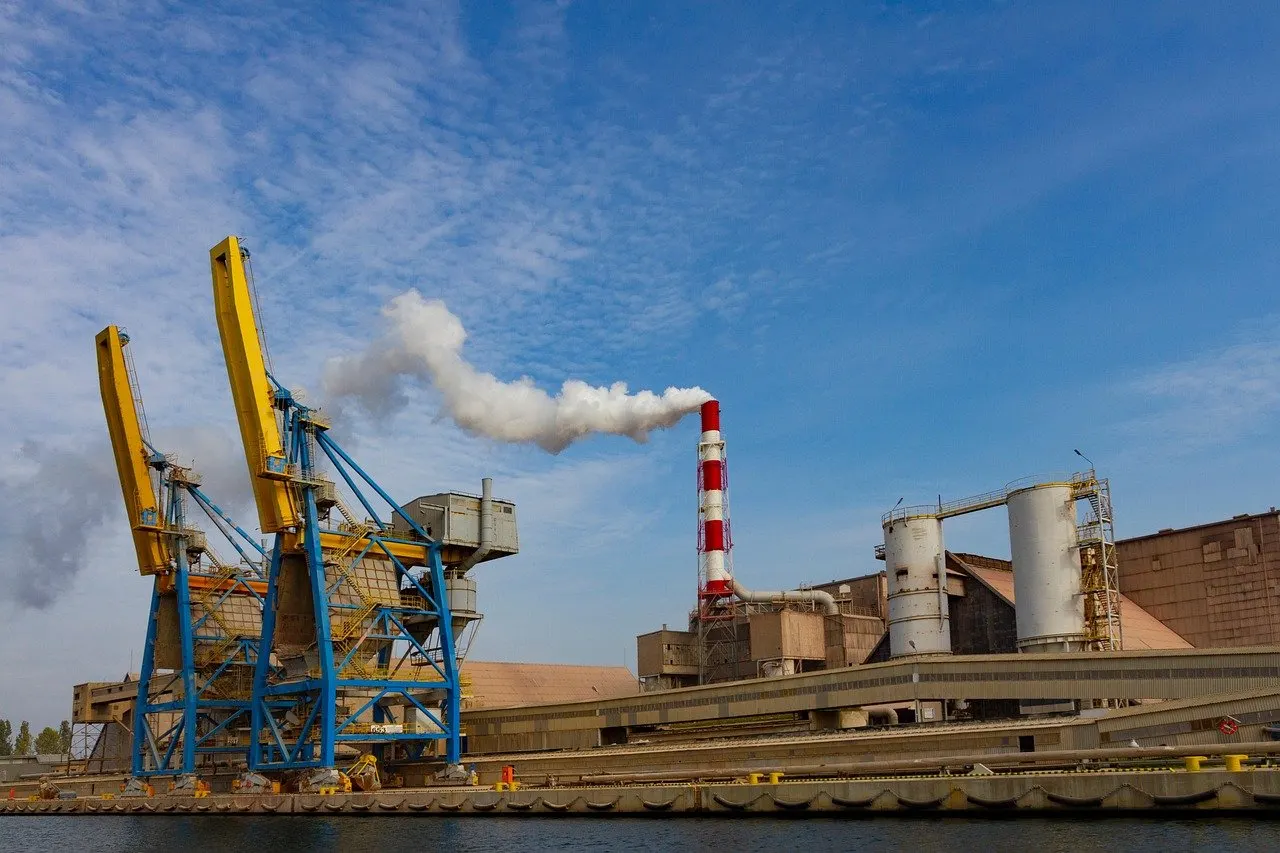 Gelb-blaue Hafenkräne neben Industrieanlage mit rot-weißem Schornstein und Rauchfahne