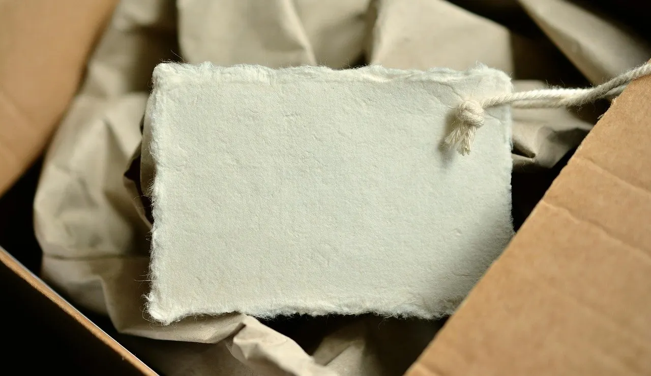 Leerer, weißer Anhänger mit Kordel liegt zwischen Packpapier in einem geöffneten braunen Karton.