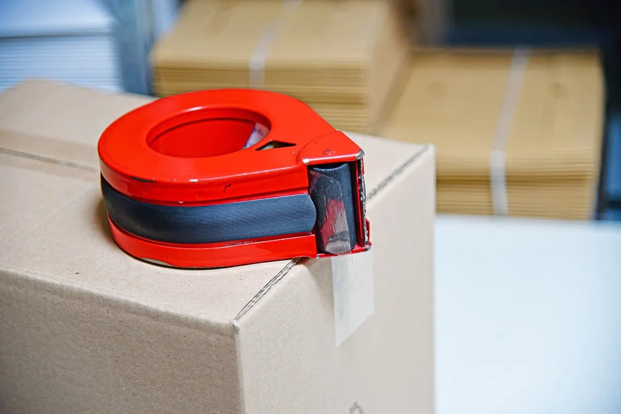 Rotes Handabroller-Gerät mit schwarzem Klebeband steht auf einem Karton, im Hintergrund weitere Kartons.