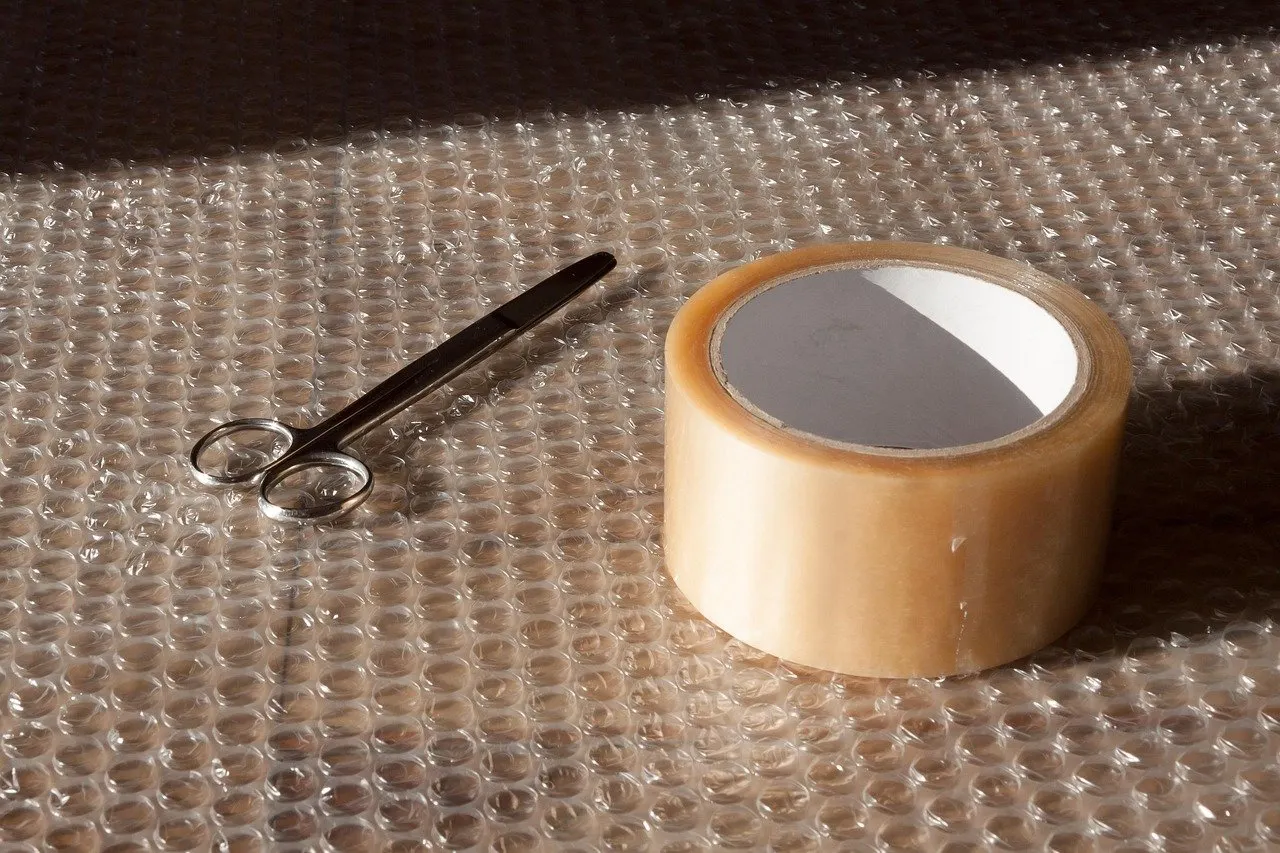 Transparentes Klebeband auf Luftpolsterfolie neben einer kleinen Schere, Sonnenlicht wirft Schatten.