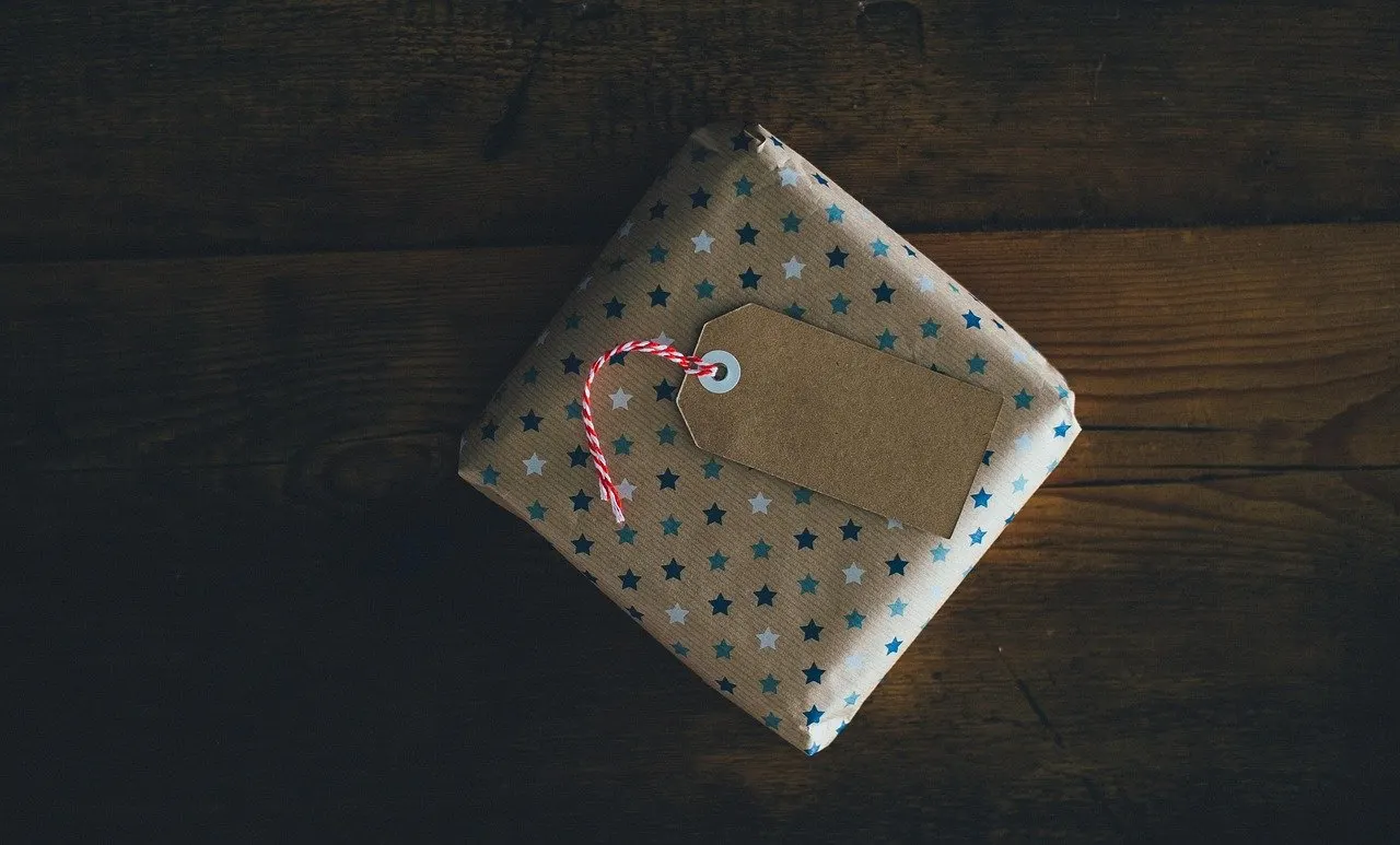 Quadratisches Paket mit Sternenmuster-Geschenkpapier, Kraftpapier-Anhänger und rot-weißer Kordel auf dunklem Holztisch.